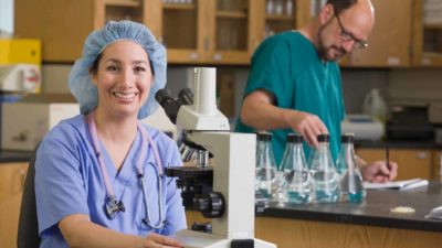 A female scientist sits at a microscope in a Universal Biosensors laboratory smiling while her colleague checks beakers of COVID-19 samples in the background.