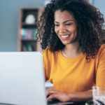 a smiling woman sits at her computer at home with a coffee alongside her, as if pleased with her investments.