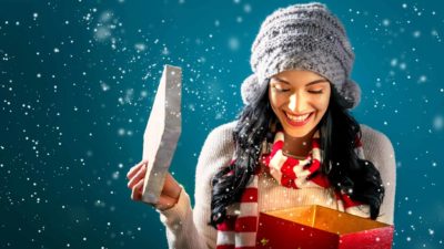 A young woman wearing a beanie as the snow falls around her smiles and opens a Christmas present in a box looking excited and smiling to represent the special dividend for Grange Resources shareholders announced today