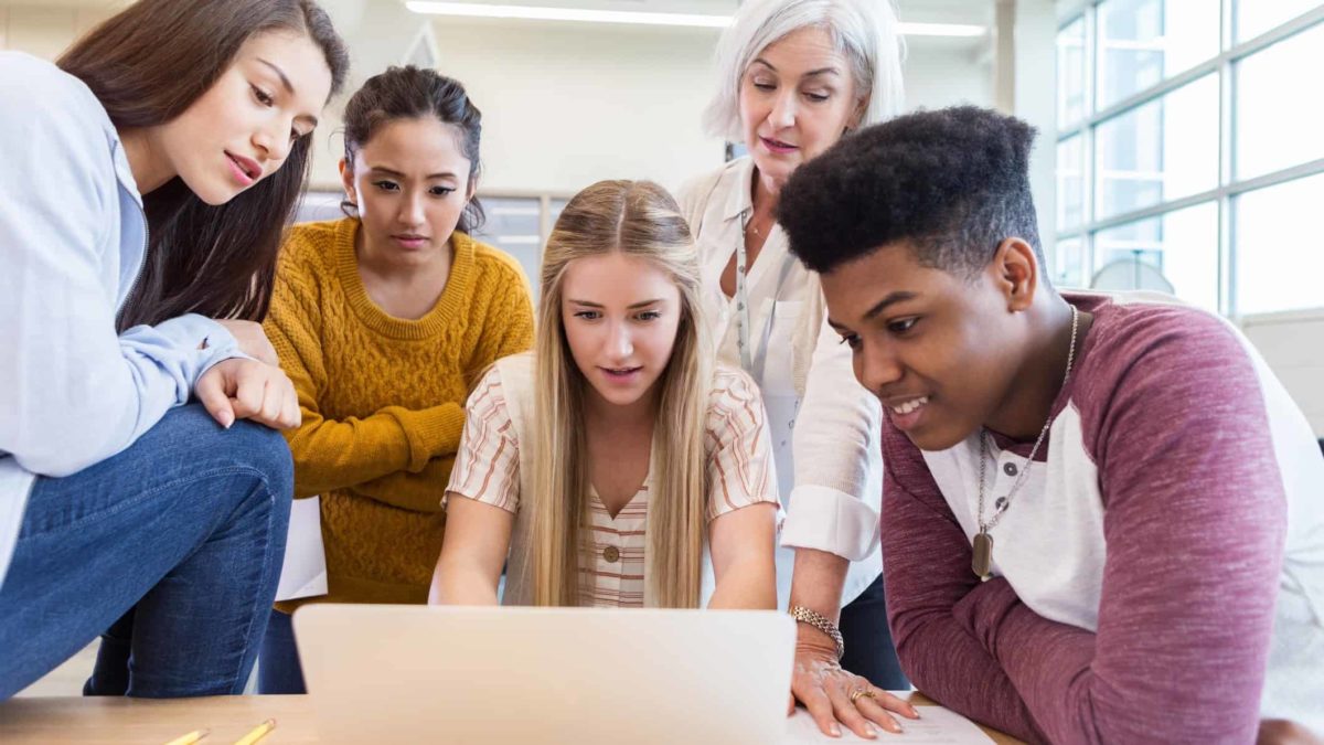 A group of young ASX investors sitting around a laptop with an older lady standing behind them explaining how investing works.