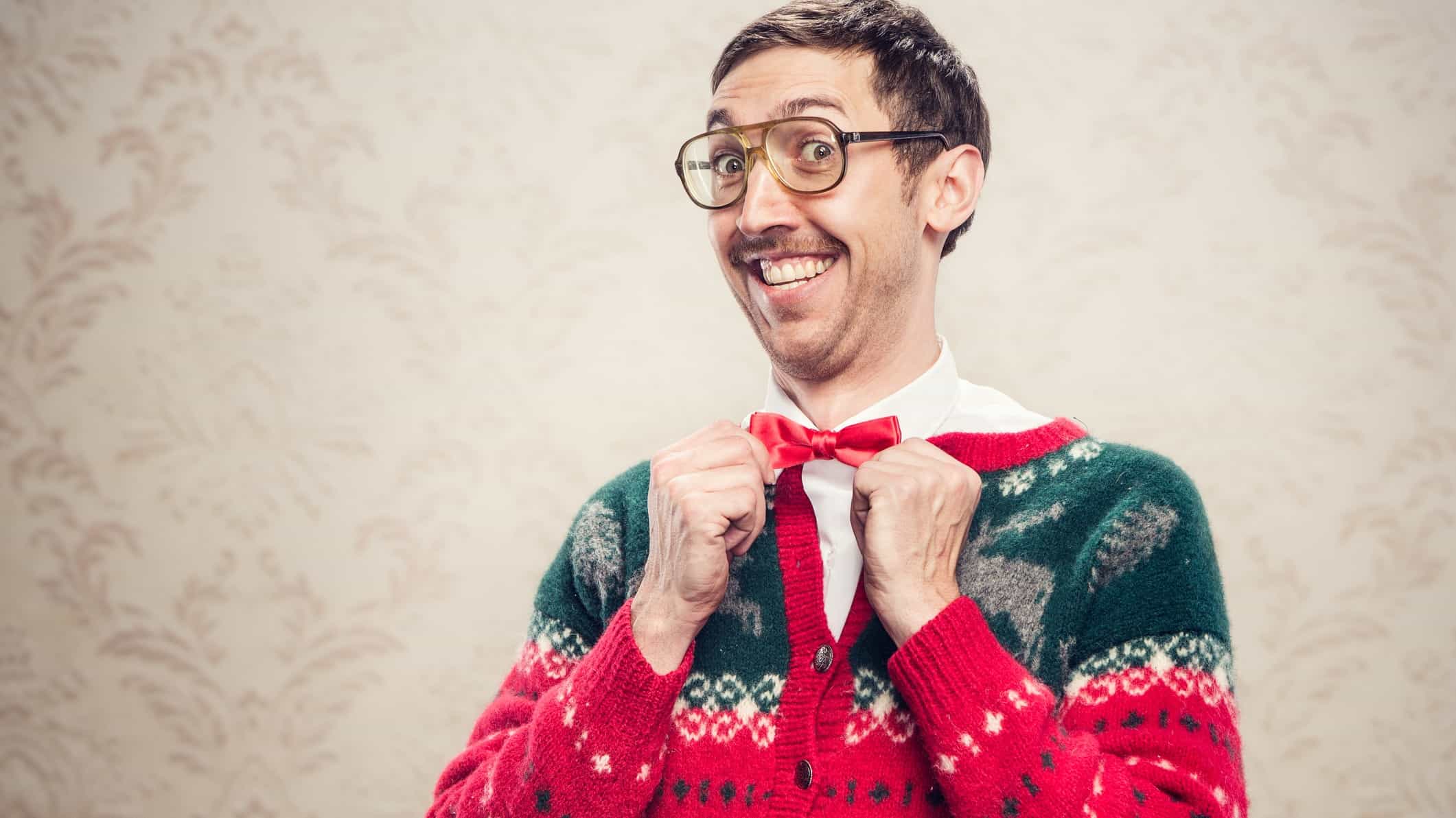 a nerdish looking man with a lovely big smile adjusts his bow tie and raises his eyebrows behind his thick glasses, as he wears a heavy knitted Christmas sweater in traditional Christmas colours of green and red.