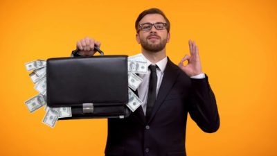 A man in suit and tie is smug about his suitcase bursting with cash.