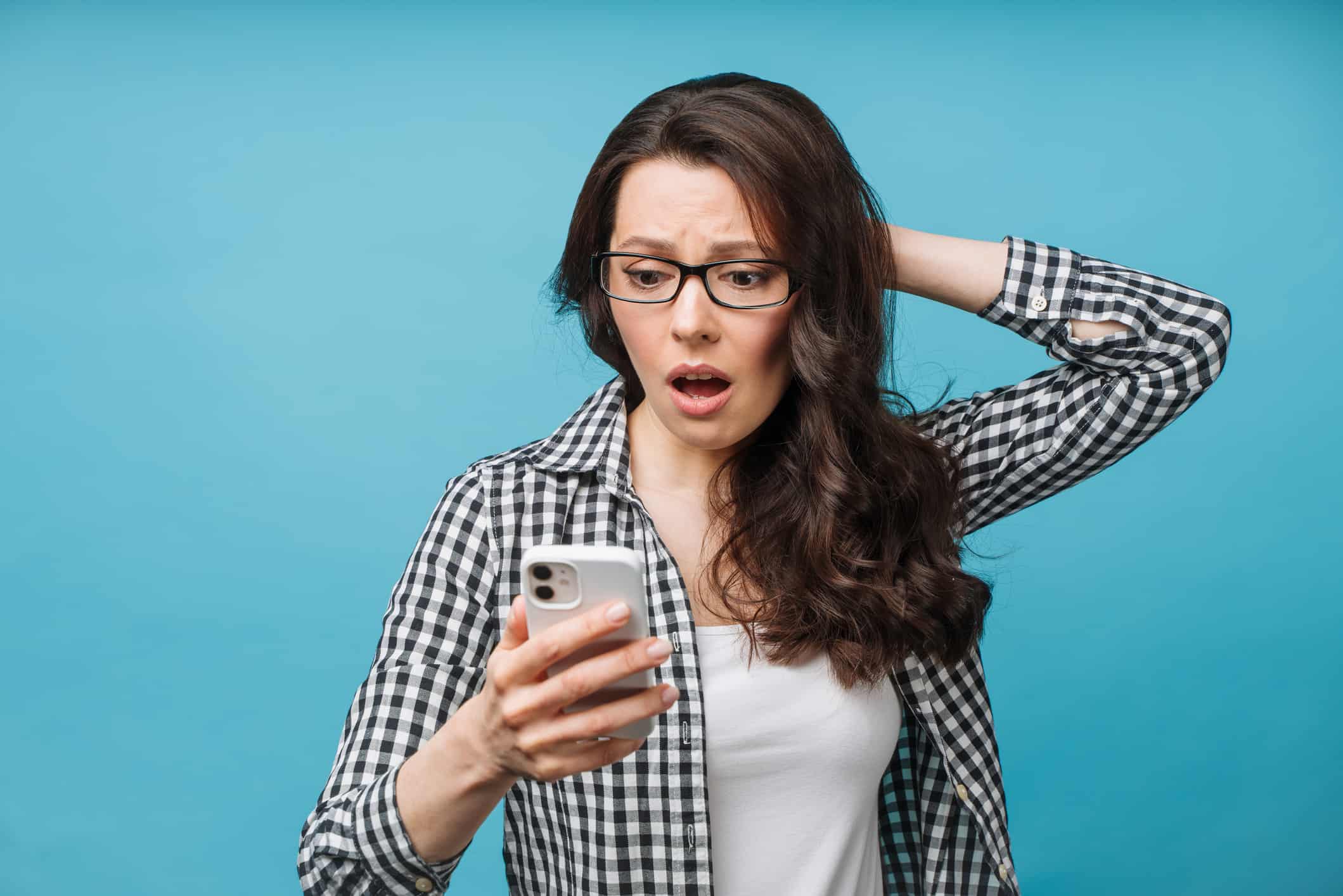 a woman looks distressed as she stares dramatically at her phone whiloe holding her hand to the back of her head with a disbelieving look on her face as though she is experiencing loss or disappointment.