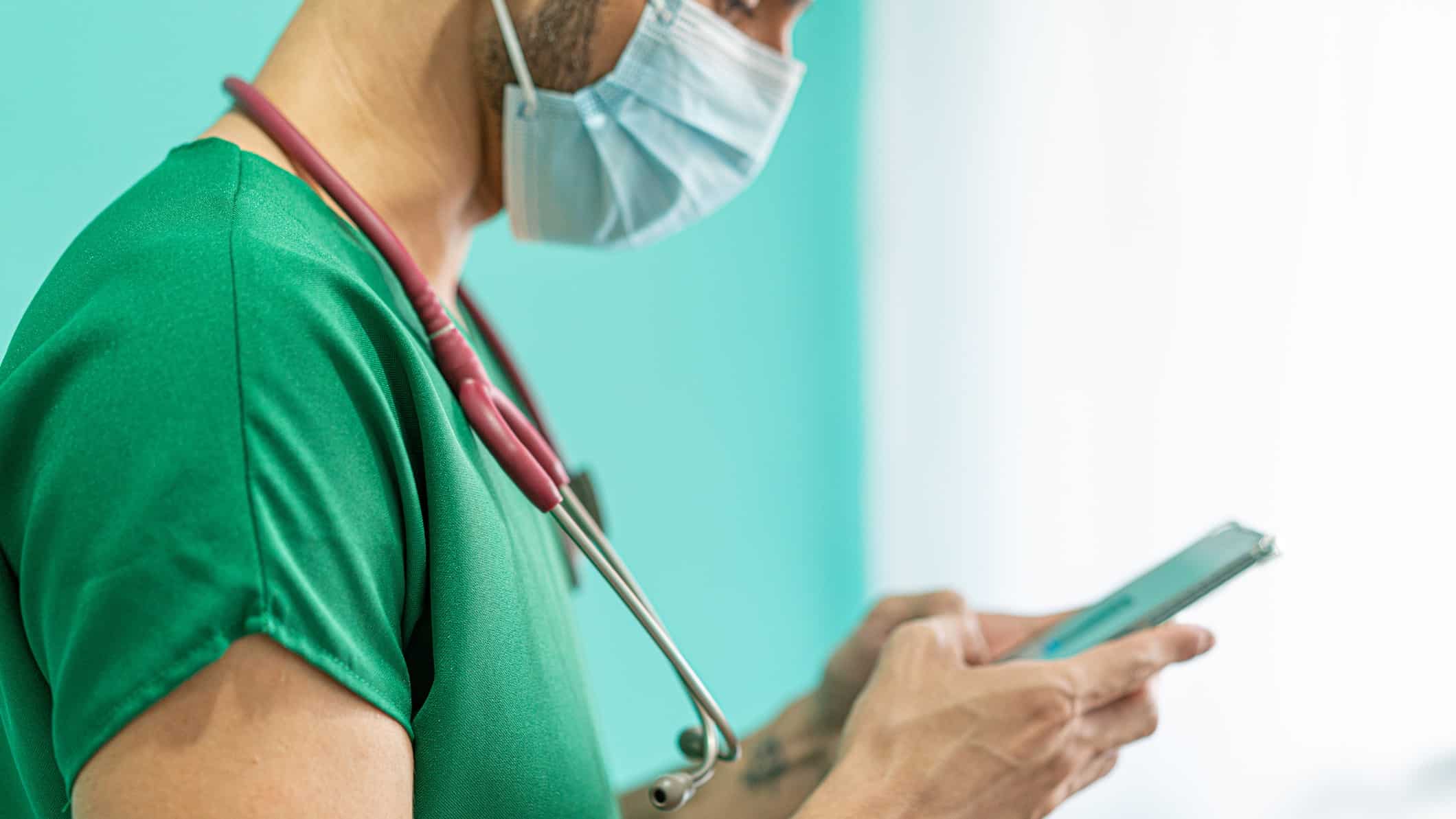 A health professional wearing green scrubs and a face mask and stethoscope uses his mobile phone.