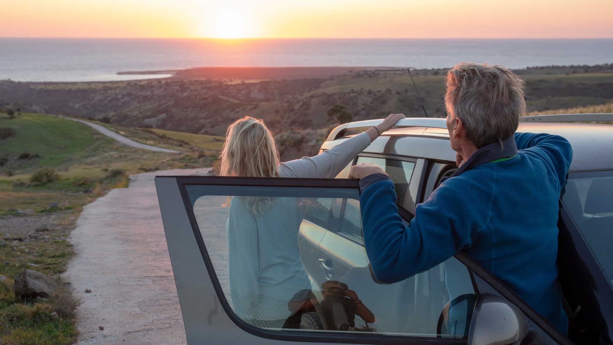 A couple hang off their car looking at the sun rising over the horizon.