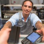 A man with long hair and tattoos holds out an EFTPOS payment machine from behind a shop counter.