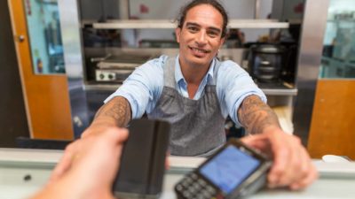 A man with long hair and tattoos holds out an EFTPOS payment machine from behind a shop counter.