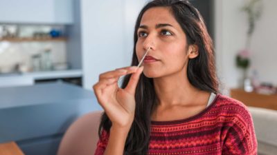 a woman inserts a swab up her nostril while conducting a rapid antigen test for COVID-19 in her home.