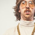 A man wearing 70s clothing and a big gold chain around his neck looks a little bit unsure.