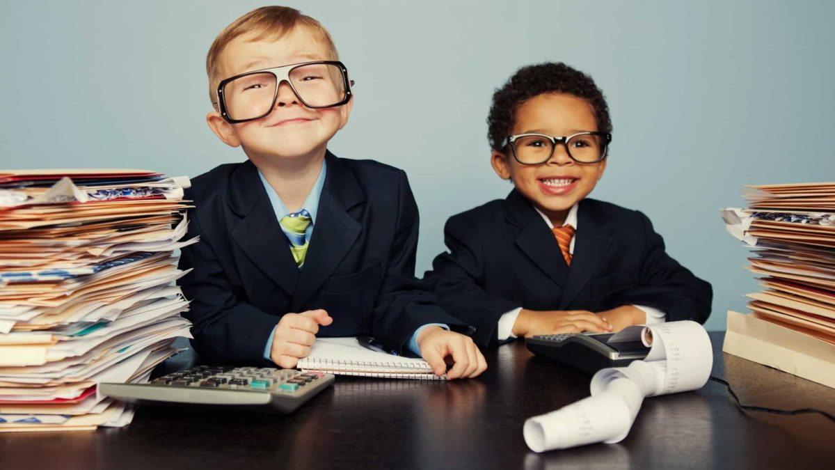 two cute young boys dressed in business suits sit amid a pile of papers with a calculator and adding machine looking very happy for themselves.
