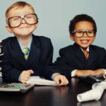 two cute young boys dressed in business suits sit amid a pile of papers with a calculator and adding machine looking very happy for themselves.