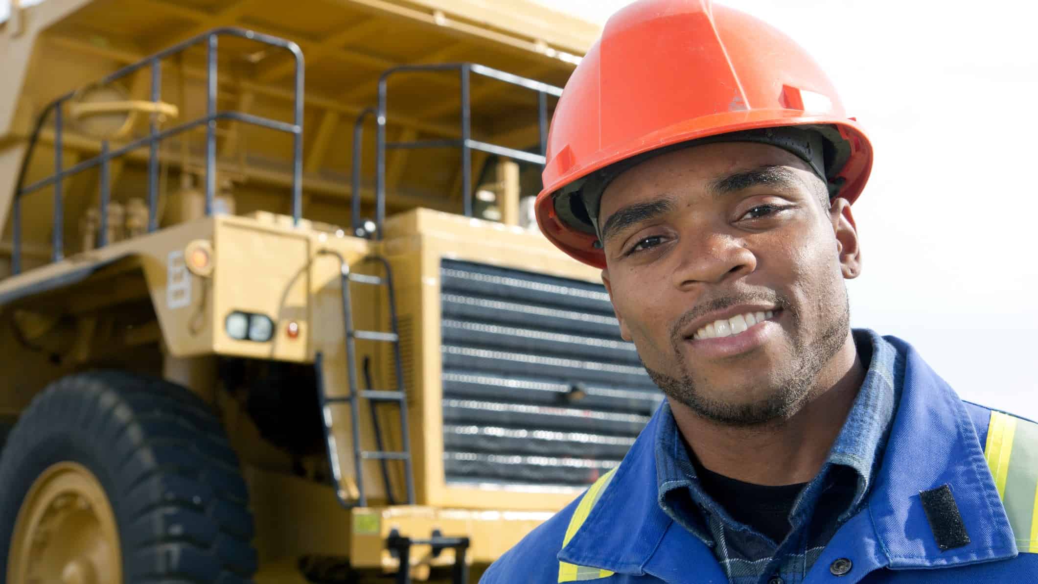 A Paladin Energy miner wearing a hard hat and protective gear stands in front of a large mining truck and smiles to the camera.