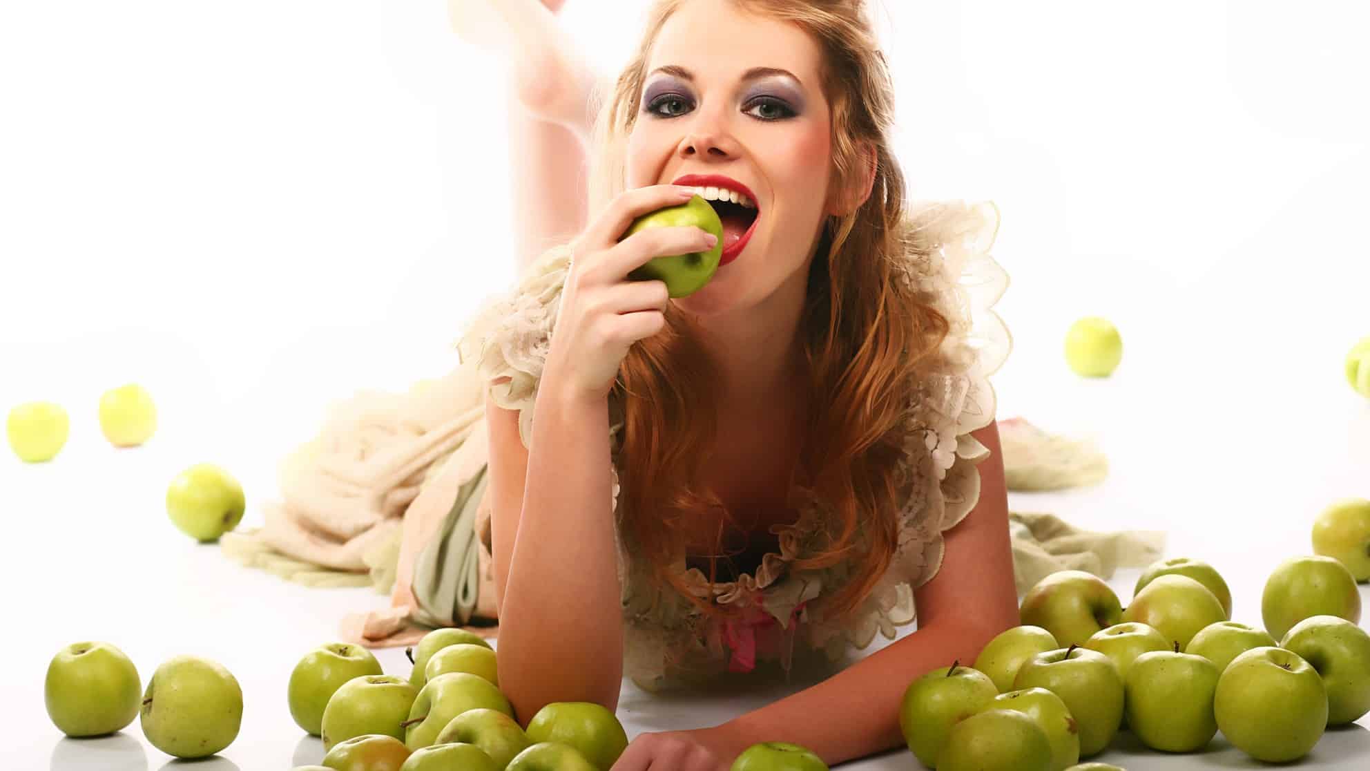 a young woman lies on the floor propped on her elbows holding a green apple to her mouth amid a large scattering of green apples around her on the floor. She is smiling and holding her mouth wide open as she is about to take a big bite of the apple she holds in her hand near her mouth.