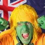 Man in green face paint and yellow wig/hat cheers in front of an Australian flag.