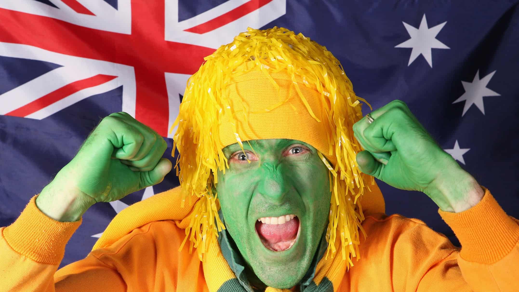 Man in green face paint and yellow wig/hat cheers in front of an Australian flag.