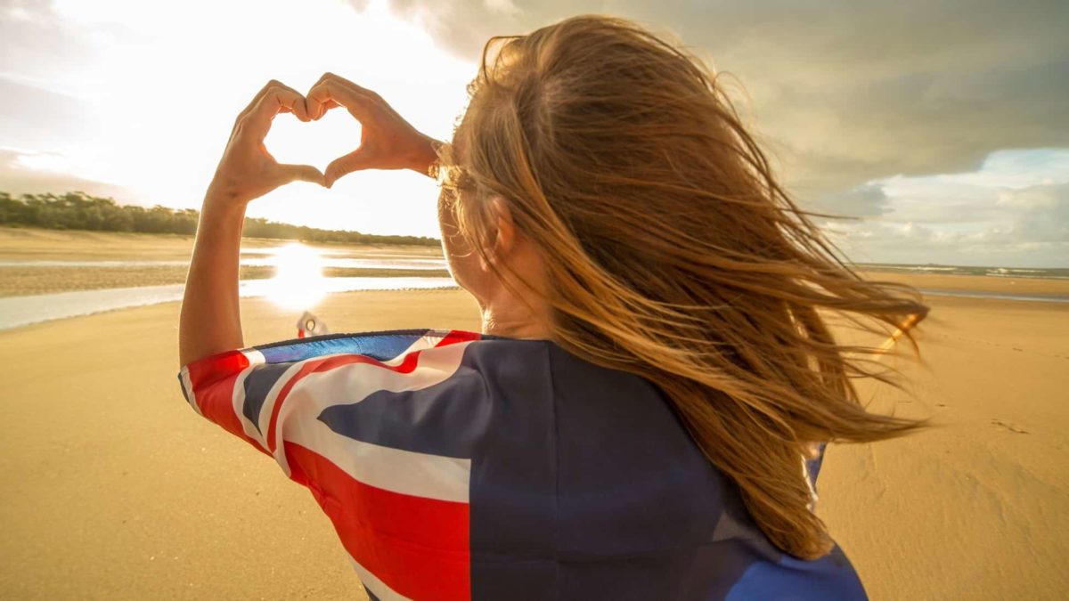 A woman faces away from the camera as she stand on the beach with an Australian flag around her shoulders and making a heart shape with her hands.