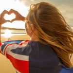 A woman faces away from the camera as she stand on the beach with an Australian flag around her shoulders and making a heart shape with her hands.