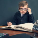 a young boy dressed in a business suit and wearing thick black glasses peers straight ahead while sitting at a heavy wooden desk with an old-fashioned calculator and adding machine while holding a pen over a large ledger book.