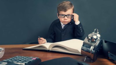 a young boy dressed in a business suit and wearing thick black glasses peers straight ahead while sitting at a heavy wooden desk with an old-fashioned calculator and adding machine while holding a pen over a large ledger book.