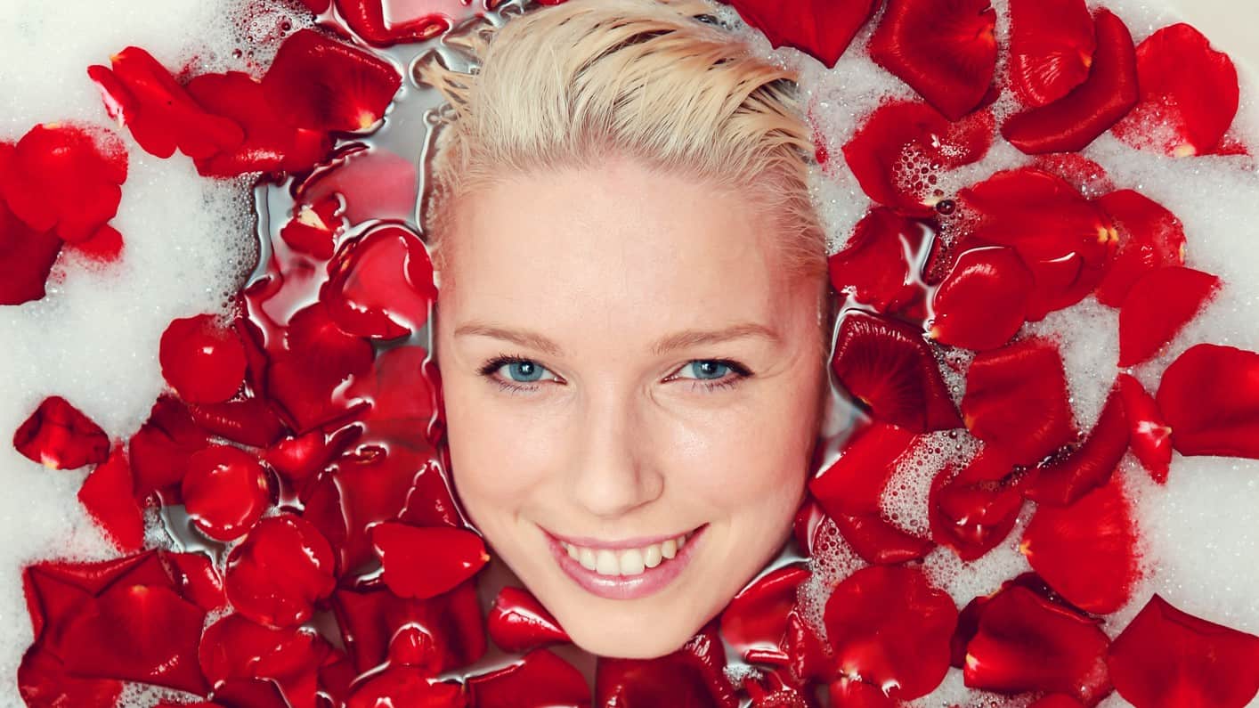 a smiling attractive woman's face appears in the midst of rose petals floating on bath water.