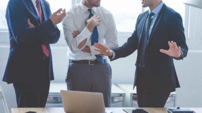 a group of 3 faceless business men stand together with one extending his hands dramatically as if protesting his treatment or stating his case passionately.