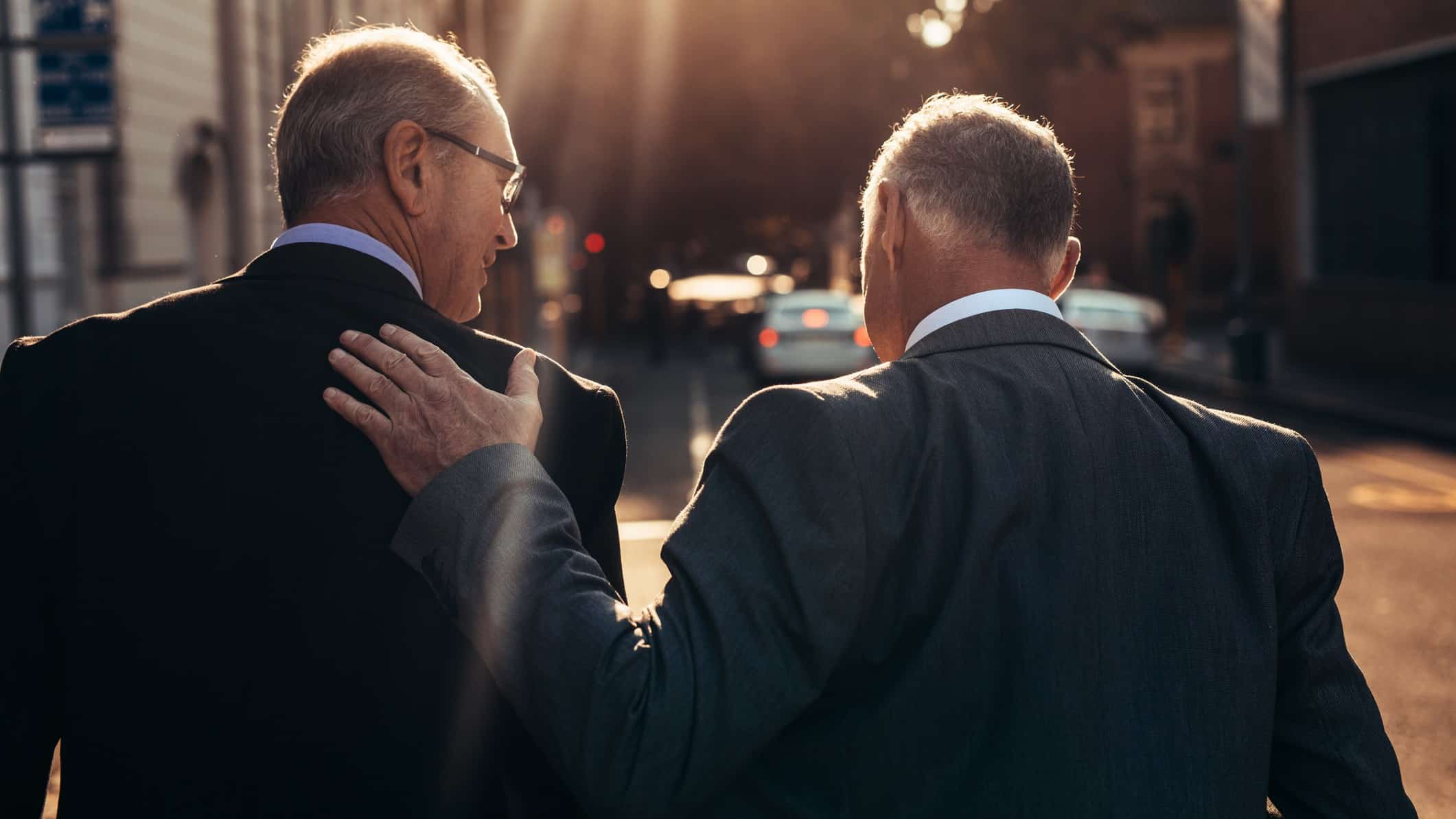 two men in suits with their backs to the camera walk off into a sunset on a city street with one placing his hand on his companion's shoulder as if in a fond gesture.