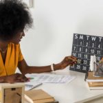 a woman sitting at a desk checks an old fashioned calendar resting against her wall as she sits with documents in front of her.