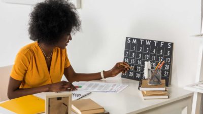 a woman sitting at a desk checks an old fashioned calendar resting against her wall as she sits with documents in front of her.