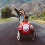 a small child and a pug dog sit in a go cart wearing old fashioned drivers headress and goggles as the drive along a country road with the boy holding his arm in the air and shouting as if celebrating their performance behind the wheel.