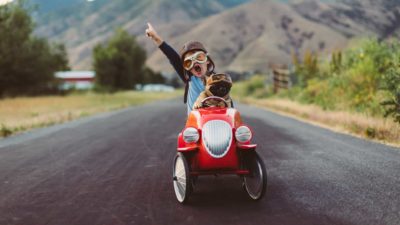 a small child and a pug dog sit in a go cart wearing old fashioned drivers headress and goggles as the drive along a country road with the boy holding his arm in the air and shouting as if celebrating their performance behind the wheel.