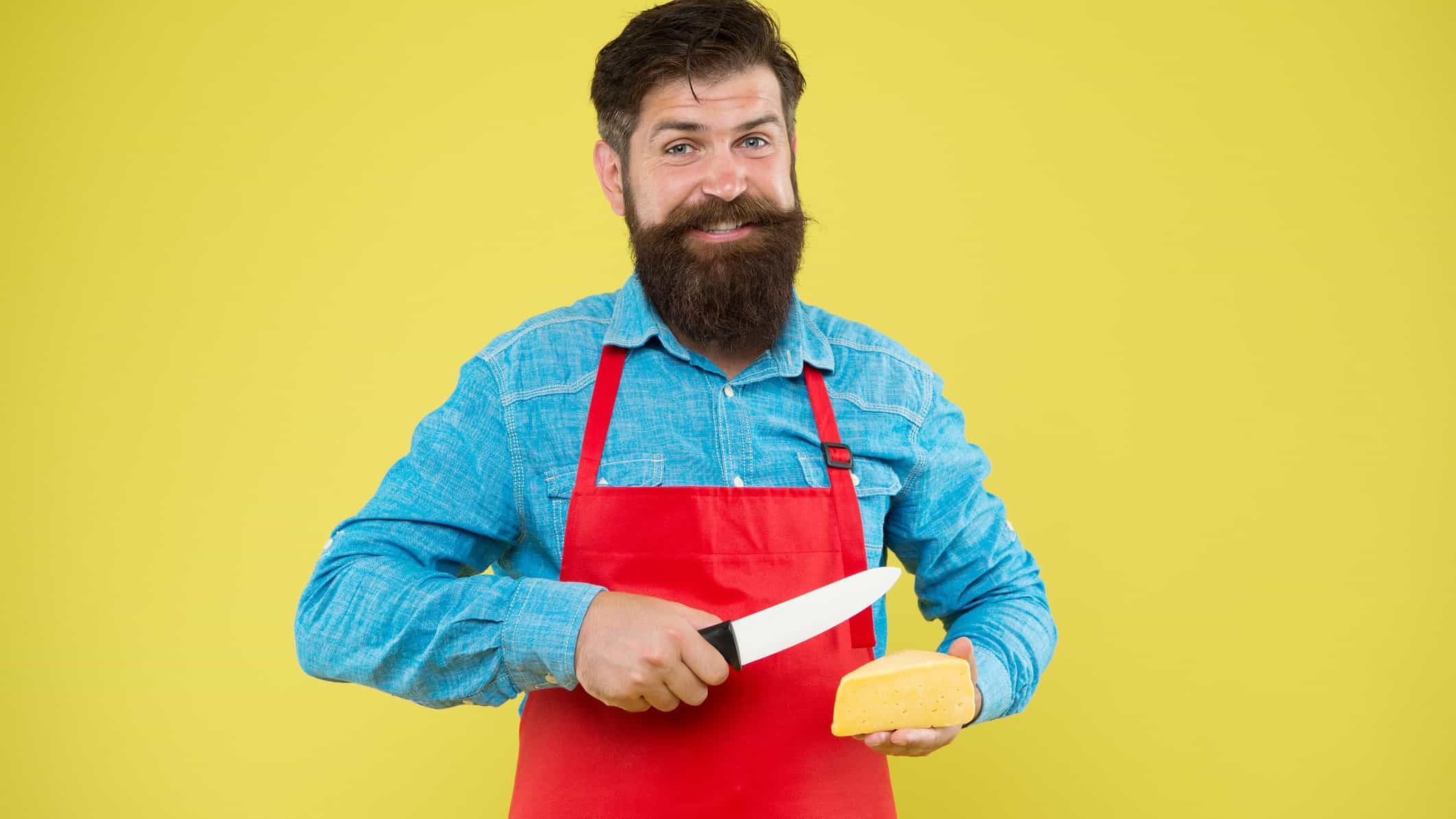 a bearded man with a big smile wearing a bright red apron holds a knife in one hand and a big slab of cheese in the other as though he is about to slice it.