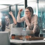 A happy group of workers around a table raise their arms in the air as though celebrating a work achievement. One woman is on her feet with her arm raised in the air in a fist-pumping action.
