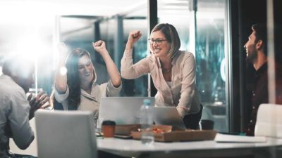 A happy group of workers around a table raise their arms in the air as though celebrating a work achievement. One woman is on her feet with her arm raised in the air in a fist-pumping action.