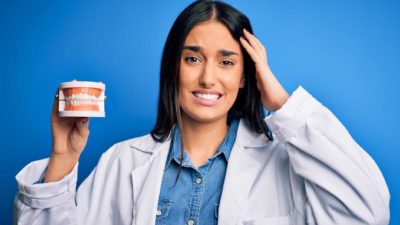 a woman wearing a white coat like a dentist holds a pair on dentures with a grimacing look on her face and her other hand to her head