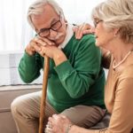 an elderly couple site together on a sofa in their home with the old man leaning forward on his walking stick and the elderly woman beside him offering comfort by resting her hand on his shoulder.