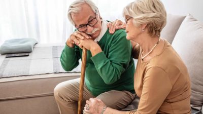 an elderly couple site together on a sofa in their home with the old man leaning forward on his walking stick and the elderly woman beside him offering comfort by resting her hand on his shoulder.