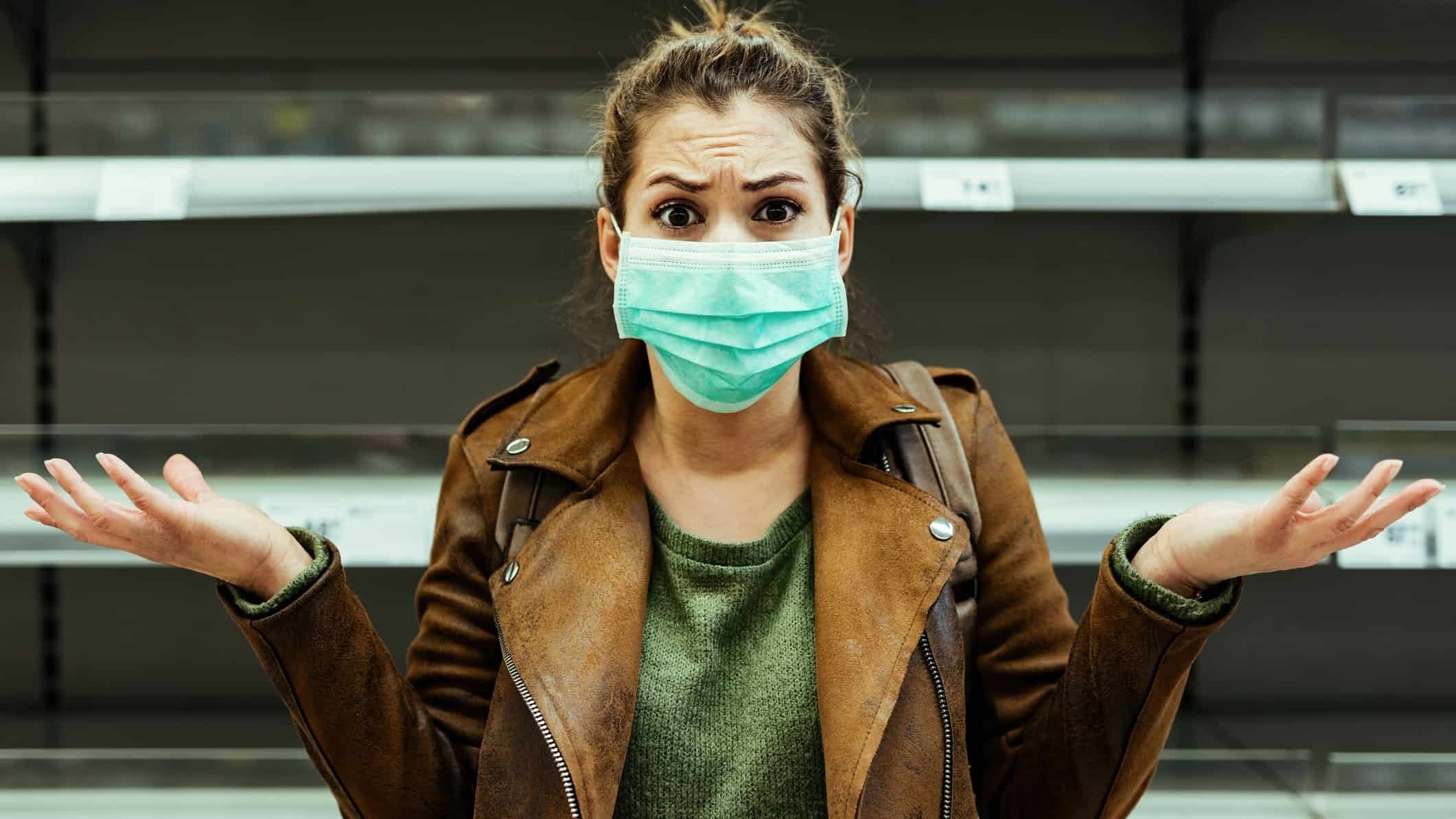 Shocked woman with protective mask gesturing while standing in front of empty shelf at supermarket during coronavirus pandemic.
