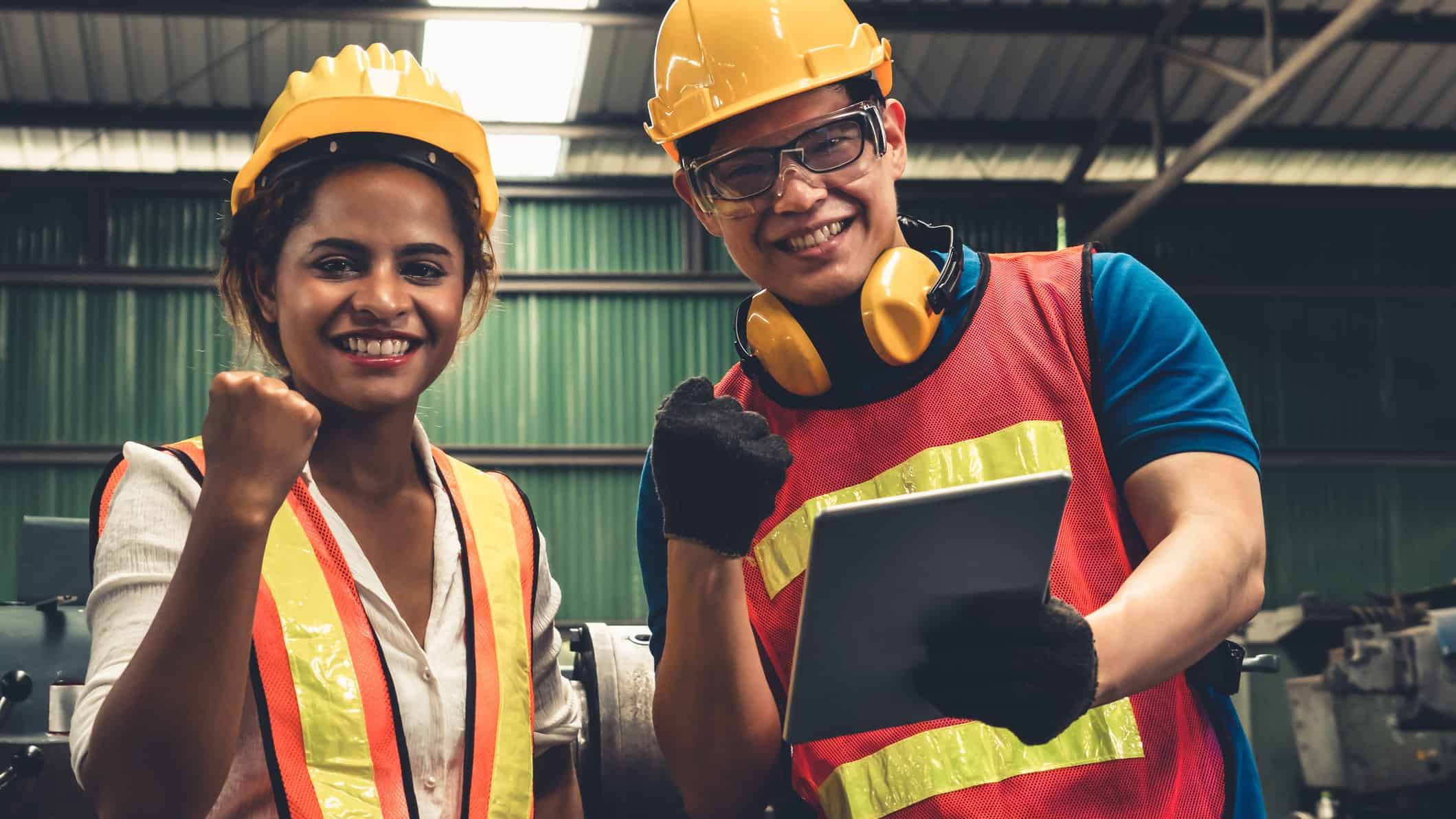 two workers in hard hats and high visibility gear give celebratory fist pumps while checking paperwork at a processing site with equipment in the background.