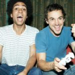 two young men sit side by side with gaming controllers pumping their fists and celebrating with joyous looks on their faces at their achievements in the video game they are playing.