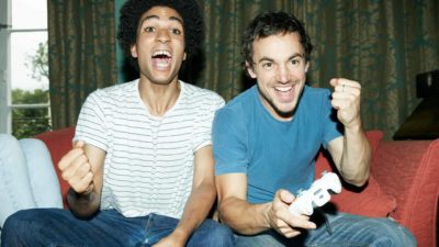 two young men sit side by side with gaming controllers pumping their fists and celebrating with joyous looks on their faces at their achievements in the video game they are playing.
