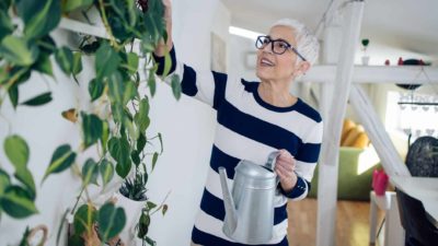 a mature but cool older woman holds a watering can and tends to a healthy green plant growing up the wall in her house.
