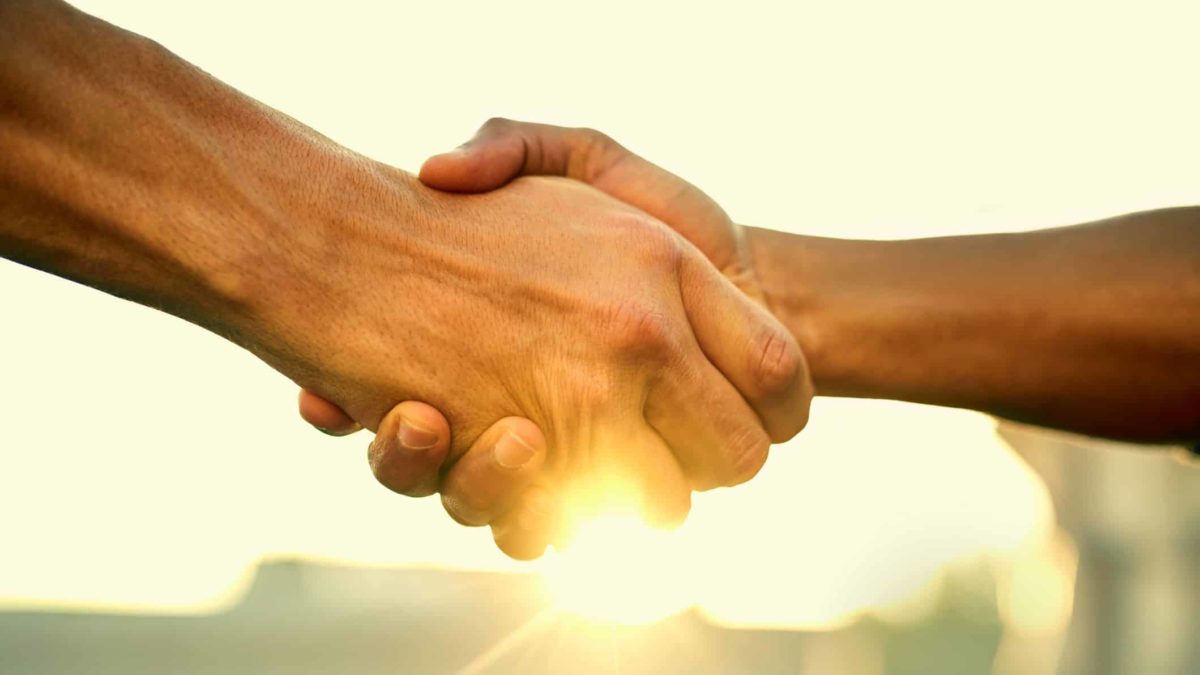 a close up of two people shake hands in front of the backdrop of a setting sun in an outdoor setting.