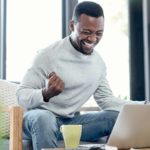 a man sits at his computer pumping his fist as he smiles widely with eyes closed and an expression of great joy as he looks at his laptop screen in his own home with a cup nearby.