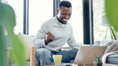 a man sits at his computer pumping his fist as he smiles widely with eyes closed and an expression of great joy as he looks at his laptop screen in his own home with a cup nearby.