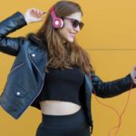 a girl wearing headphones strikes a dance pose as she smiles at her phone being held in her hand as if a great song is being played through her music setup.
