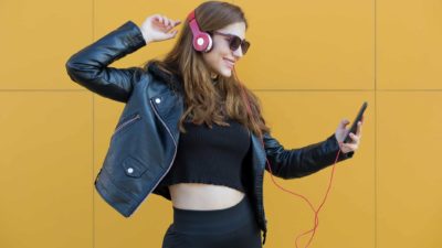 a girl wearing headphones strikes a dance pose as she smiles at her phone being held in her hand as if a great song is being played through her music setup.