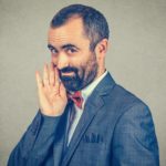 A man in a business suit holds his hand up to his mouth as though sharing a secret and gives a sly grin.