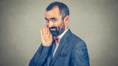 a man in a business suit holds his hand up to his mouth as though sharing a secret and gives a sly grin.