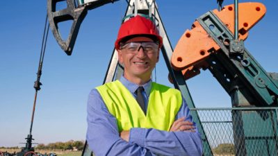 An oil refinery worker stands in front of an oil rig with his arms crossed and a smile on his face as the Woodside share price climbs today