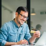 Happy man wearing a blue shirt and glasses holding a card and using buy now pay later services to purchase a product on his office computer